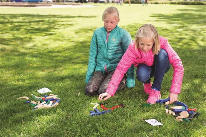 Two girls wearing bright jackets sit and kneel on grass in a park, playing with the Buitenspeel Sport Blocks Puzzle Game arranged in small groups. Sunlight filters through trees, casting dappled shadows on the lawn.