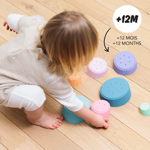 A child is playing with a Stak Baby Toy, enhancing their fine-motor skills, on a wooden floor.