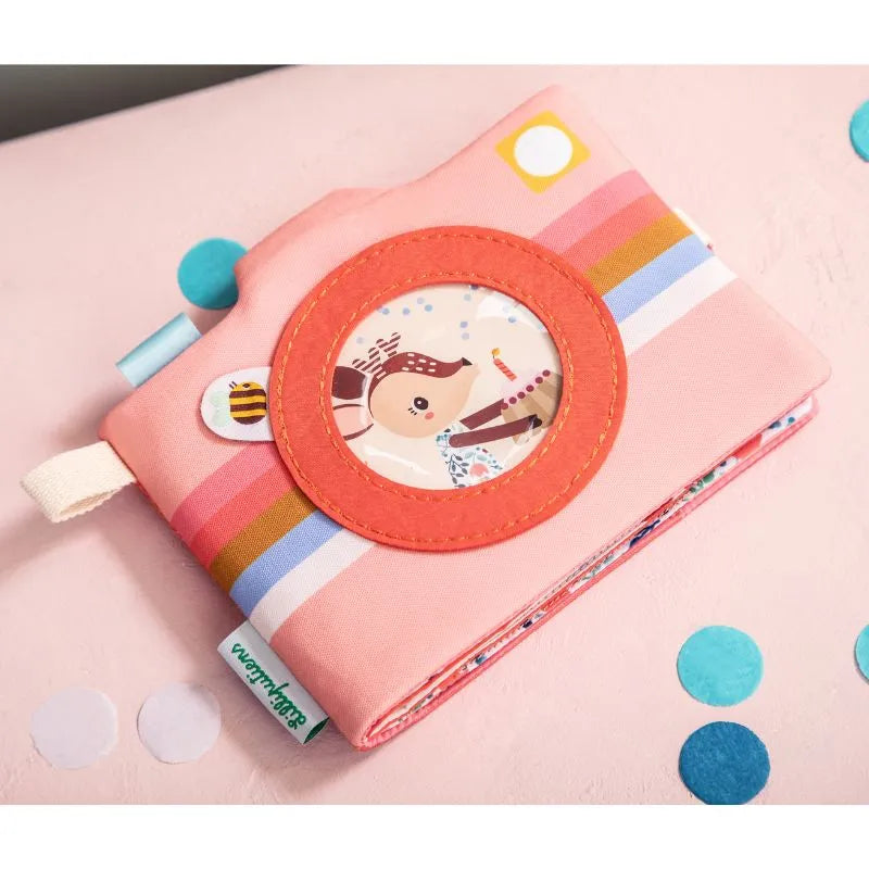 A pink fabric toy camera with colorful stripes and an animal illustration lens rests on a soft pink surface. Blue and teal confetti scattered around evoke memories of baby's first moments, like those in the Lilliputiens I'm Discovering Stella Photo Album, after a fun car journey.