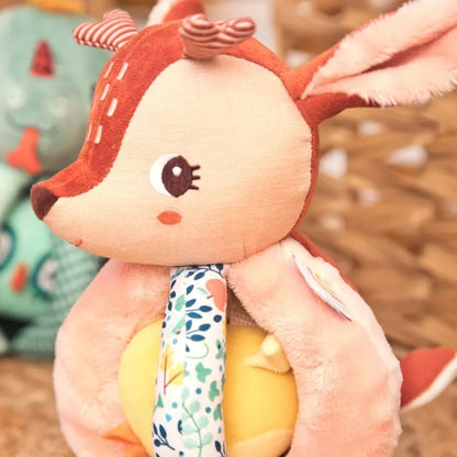 The Lilliputiens "I'm Discovering Stella The Faon" rattle features a plush deer design with large ears, embroidered details, textured handles, and a floral-patterned neck over its yellow body. The blurred background suggests another toy nearby.