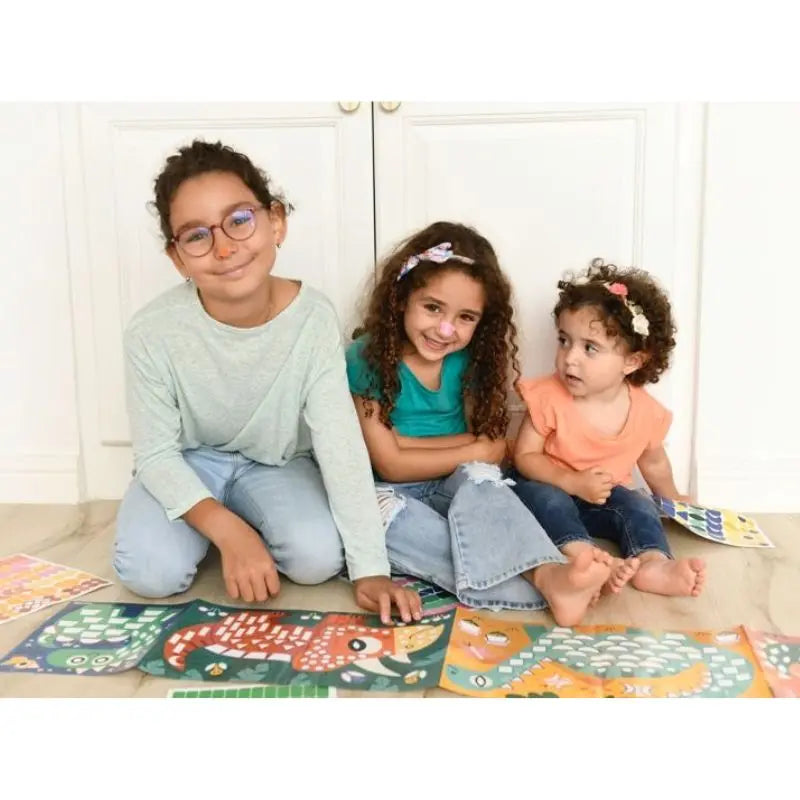 Three young children sit on the floor indoors, smiling and enjoying the Poppik Sticker Panorama – Dinosaurs—a fun and creative board activity for kids. They wear casual clothes and are in front of white cabinets.