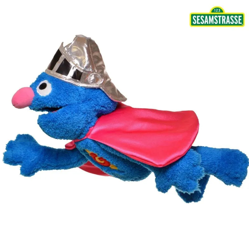 A Living Puppets Super Grover Hand Puppet, featuring a plush blue character with a red cape and silver helmet, is flying. The cheerful figure with a pink nose perfectly embodies the spirit of Sesame Street. A logo in the top right corner reads "Sesamstrasse.