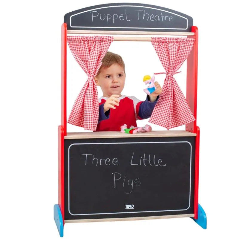 A child is standing behind the Tidlo Wooden Puppet Theatre, which features red and white checkered curtains. The theatre's sign reads "Puppet Theatre," and the blackboard below announces "Three Little Pigs." Engaged in imaginative, creative play, the child is holding a hand puppet and performing a delightful show.