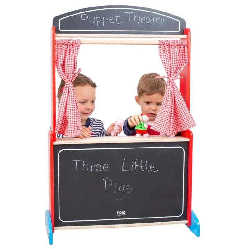 Two children are engaged in creative play with hand puppets behind a small Tidlo Wooden Puppet Theatre adorned with red gingham curtains. The top reads "Puppet Theatre," and the chalkboard front panel displays "Three Little Pigs," making this delightful children's toy a perfect backdrop for their imaginative stories.