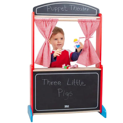A young child stands behind the Tidlo Wooden Puppet Theatre with red gingham curtains, holding puppets. The chalkboard reads "Three Little Pigs." This charming toy inspires imaginative play and creativity. A sign above says "Puppet Theatre.