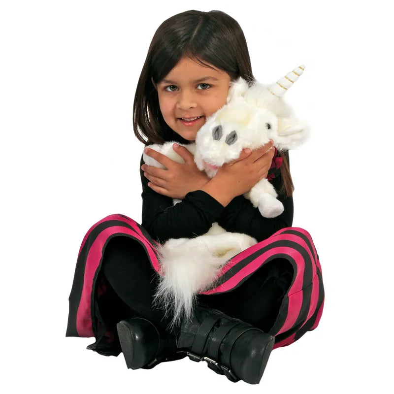 A young girl with long dark hair sits cross-legged, wearing a black outfit with pink stripes and black boots. She is hugging a white plush unicorn with a small horn and smiling at the camera, while beside her rests The Puppet Company Long Sleeved Puppet Unicorn.