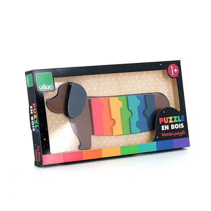 A charming Vilac Dog Wooden Puzzle, featuring rainbow-colored wooden pieces in the shape of a dachshund, is displayed in a dotted rectangular box. "Vilac" adorns the top left corner, while "Puzzle en Bois" and "Wooden Puzzle" grace the bottom right. This delightful toy is suitable for ages 1 year and up.