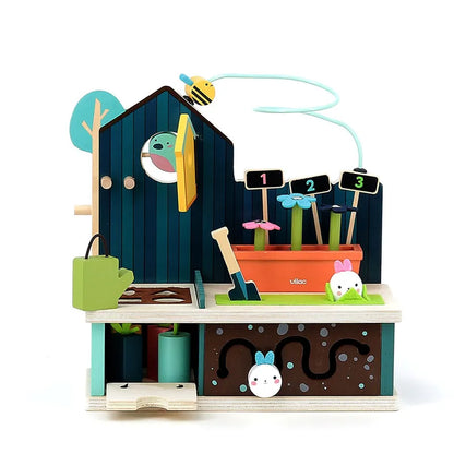 Introducing the Vilac Early Learning Garden, a vibrant wooden toy set illustrating a charming garden scene, ideal for children 18 months and older. This engaging set includes a house-like structure with a moveable bird, raised garden beds numbered one to three, a bee on a wire hovering above, and various gardening accessories such as a shovel and hanging plants.