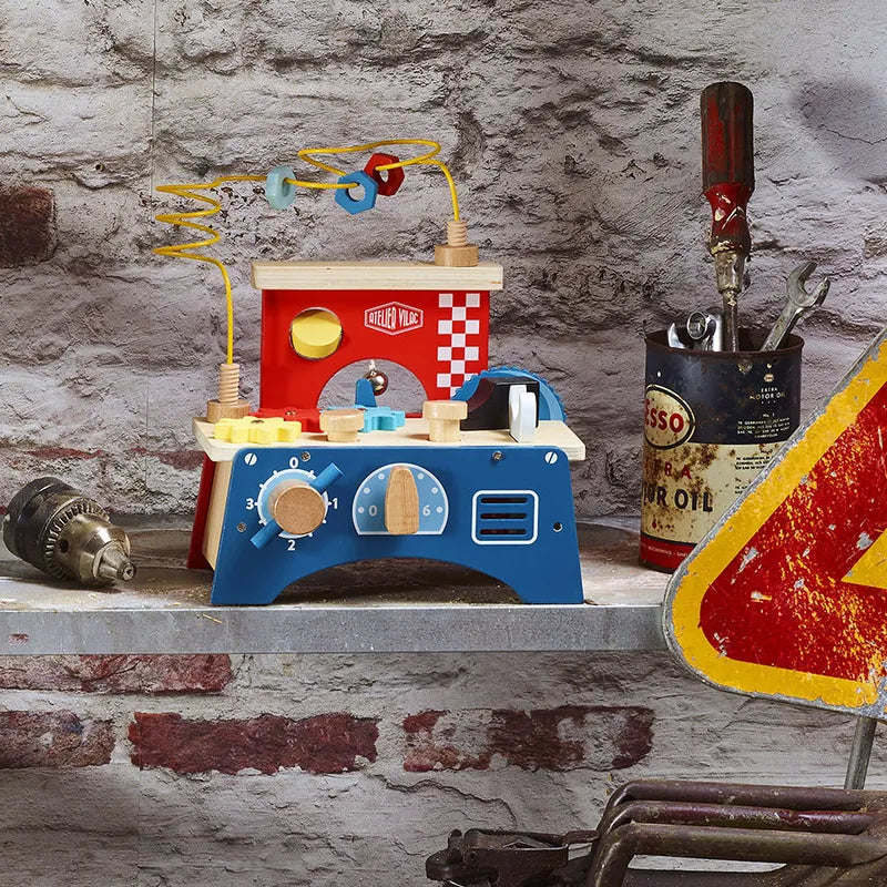 The Vilac Early Learning Workbench, featuring colorful knobs and dials, is placed on a countertop against a rustic brick wall. Beside it, there are metal cans with paintbrushes and tools, including a drill and a triangular road sign with a red border, creating a playful setting suitable for kids 18 months and older.