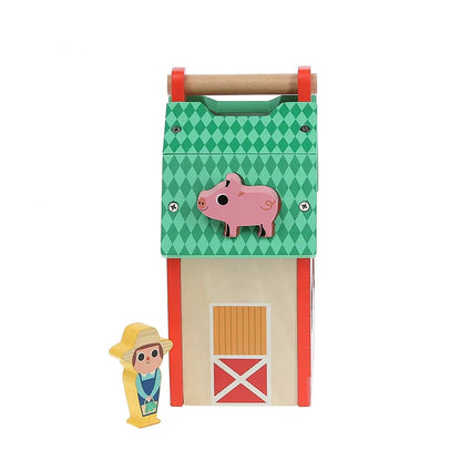 The Vilac My Nice Farm Shape Sorter features a wooden toy barn with a green and red diamond-patterned roof and a pink pig decoration that stands upright. Beside it, there's a colorful figure of a person wearing a yellow hat and blue overalls, holding a small watering can. Ideal for toddlers aged 2 years and up, the barn includes a small door at the bottom with an X design.