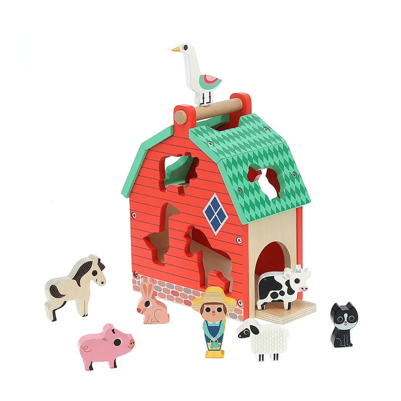 The Vilac My Nice Farm Shape Sorter is shown with a charming wooden toy barn featuring a red facade and a green roof. The set comes with various animal-shaped cutouts and wooden figures, including a horse, pig, goat, cow, sheep, goose, two rabbits, dog, and a person. Suitable for toddlers aged 2 years and up. A goose figure perches on top of the barn.