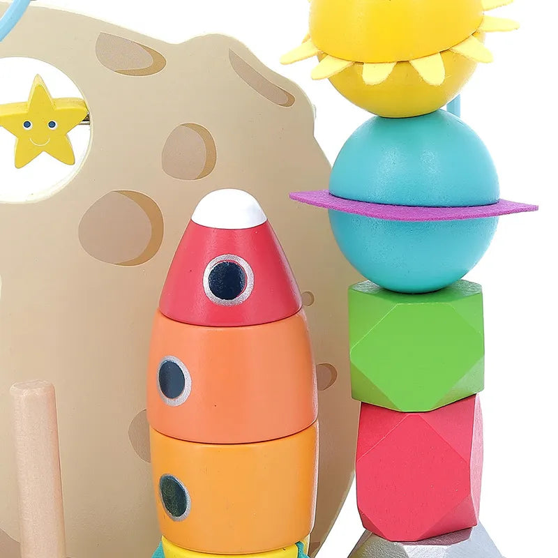 The Vilac In the Stars Galaxy Counting Game includes vibrant wooden stacking pieces, featuring a red and orange rocket ship, a sun with yellow rays, and various geometric shapes in teal, blue, green, and pink. The beige background is adorned with star and moon cutouts. These space-themed pieces are designed to be stacked in various configurations to promote creative play and early learning.