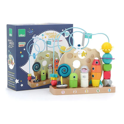 A vibrant children's wooden toy set called "Vilac In the Stars Galaxy Counting Game" is designed with a space theme. It includes a wooden board with numbered pegs (1-5), colorful beads on two looping wire tracks, and stacking blocks shaped like rockets and astronauts. The toy box in the background displays the assembled early learning toy alongside the brand name "Vilac.