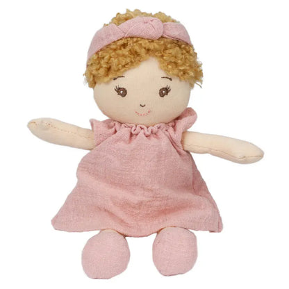 The Wilbery Baby Doll Girl Pink Outfit features soft fabric, curly blond hair, a pink dress with a matching headband, and simple stitched facial features with a sweet smile and outstretched arms.