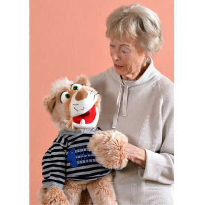 An older woman with short gray hair and a light sweater smiles while holding Wisky the large Cat Hand Puppet – 63cm, dressed in a striped shirt, against a peach-colored background.