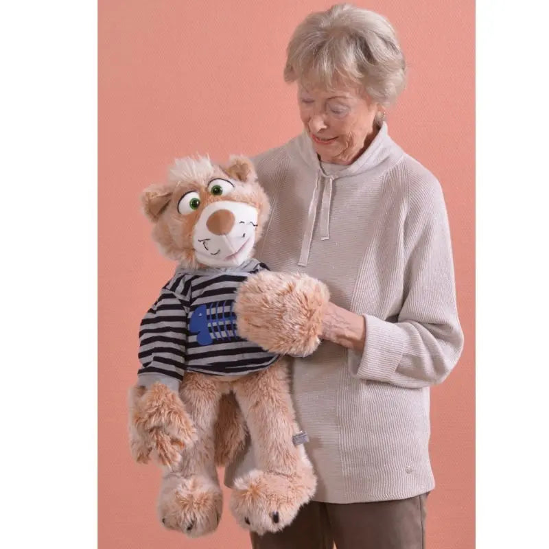 An elderly woman with short gray hair smiles while holding Wisky the large Cat Hand Puppet – 63cm, dressed in a striped shirt, standing before a plain peach-colored background.
