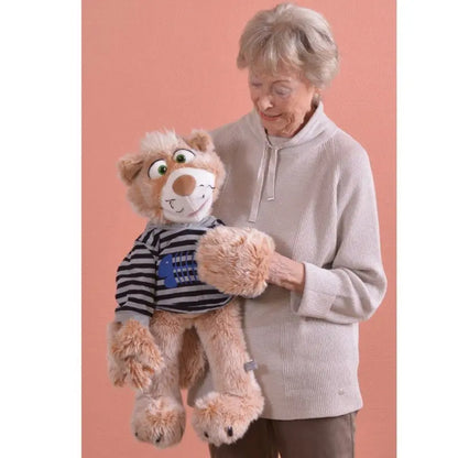 An elderly woman with short gray hair smiles while holding Wisky the large Cat Hand Puppet – 63cm, dressed in a striped shirt, standing before a plain peach-colored background.