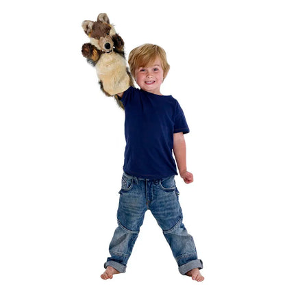 A young boy wearing jeans and a navy blue long-sleeved shirt is smiling as he uses his right hand to hold up The Puppet Company Long Sleeved Puppet Wolf, against a plain white background.