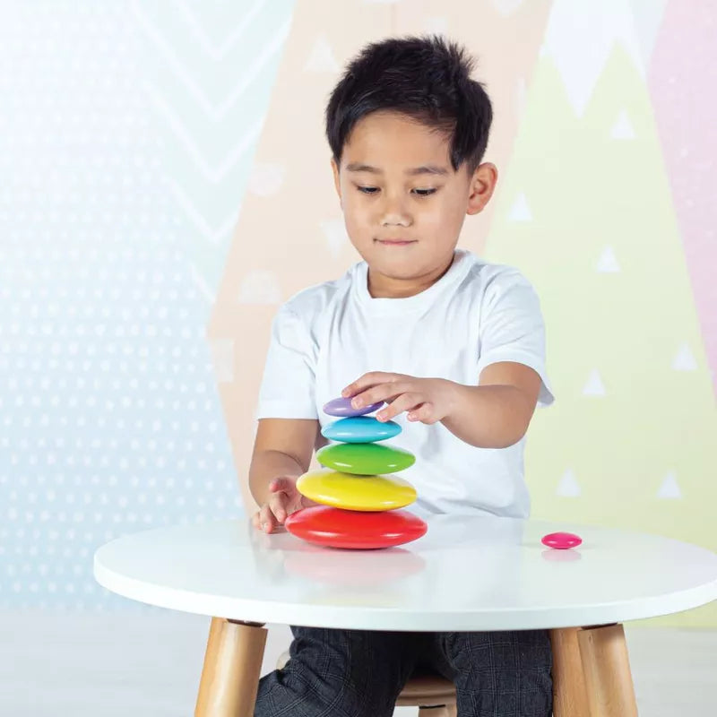 A little boy sitting at a table playing with Bigjigs Rainbow Stacking Pebbles.