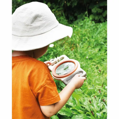 A young child in an orange shirt and white sunhat examines nature closely through the XXL Magnifying Glass “Discover”, with green foliage in the background.