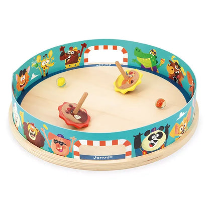 A Janod Applepop - Spinning Top Arena and children's toy with a circular wooden base and a vibrant border featuring various cartoon animals. Two spinning tops and a few marbles are inside the playing area. The sides have cut-out handles for easy carrying.