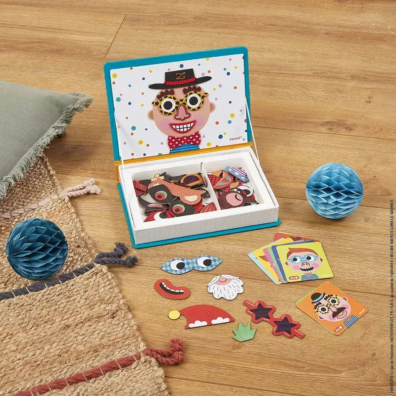 A children's magnetic toy set titled "Janod Boy's Crazy Faces Magneti'book," featuring a box with magnetic face pieces on the lid, is displayed. Various facial feature magnets and colorful cards from the Janod Magneti'books collection are spread out beside the box. The scene is set on a wooden floor with two small blue paper ball decorations nearby.