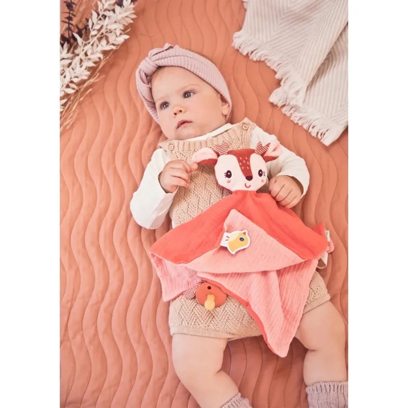 A baby wearing a beige knitted romper and a pink headband lies on a peach-colored, textured blanket. The baby holds a Lilliputiens Stella the Fawn Comforter, making it the perfect birth gift.