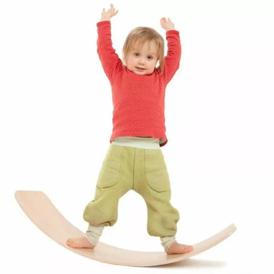A toddler in a red sweater and green pants joyfully balances on a das.Brett Bouncy Wooden Balance Board Lacquered with Cork Bottom-side Cladding, raising their hands high in the air.
