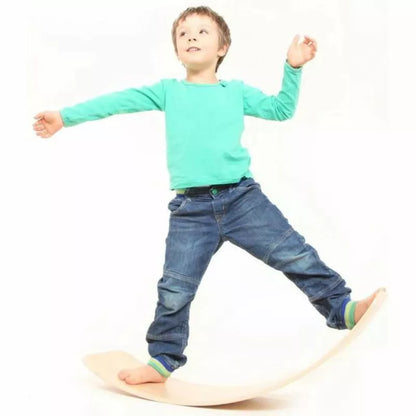 A young boy in jeans and a teal shirt joyfully balances on a das.Brett Bouncy Wooden Balance Board Lacquered with Cork Bottom-side Cladding, arms outstretched and looking upwards, on a white background.