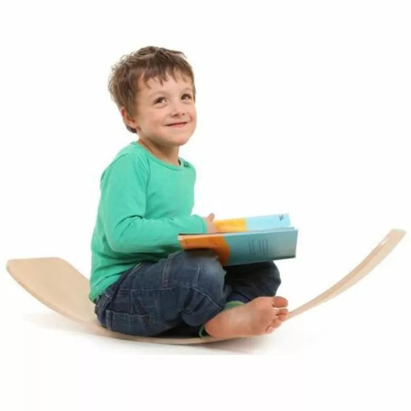 A young boy with a joyful expression sits cross-legged on a das.Brett Bouncy Wooden Balance Board Lacquered with Cork Bottom-side Cladding, holding colorful blocks. He wears a green shirt and blue jeans on a white background.