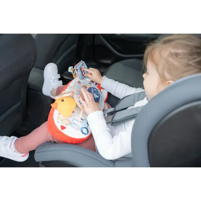 A toddler is sitting in a car seat with a safety harness on, engaged with the Lilliputiens Farm Car Activity Panel on their lap. This colorful baby toy features a yellow duck and various interactive elements. The child is wearing white shoes, pink pants, and a white long-sleeve shirt.