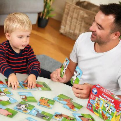 A man and a young child sit at a table playing a matching card game featuring animal images. The child is focused on the cards, while the man watches the child with a smile. A box labeled "Orchard Toys Farmyard Heads & Tails" is nearby.