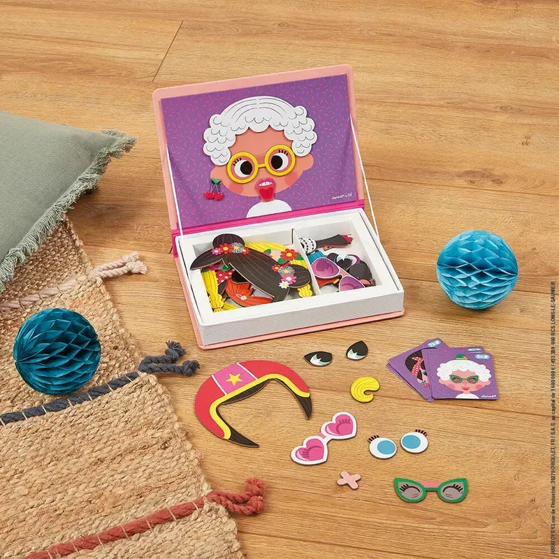 The Janod Girl's Crazy Faces Magneti'book dress-up set, featuring a box laying open on a mat, reveals various costume pieces such as a superhero mask, sunglasses, hats, eyes, and headpieces. The box lid showcases a cartoon character in glasses and lipstick. Two blue honeycomb decorations adorn the mat. Perfect as an educational magnetic game similar to other Janod Magneti'books.