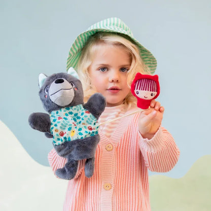 A young child with blonde hair wears a green bucket hat and a pink cardigan. The child holds a stuffed gray wolf toy in one hand and a small red doll resembling Lilliputiens Louis Wolf Handpuppet in the other, standing against a light blue background, ready for imaginative and creative play.