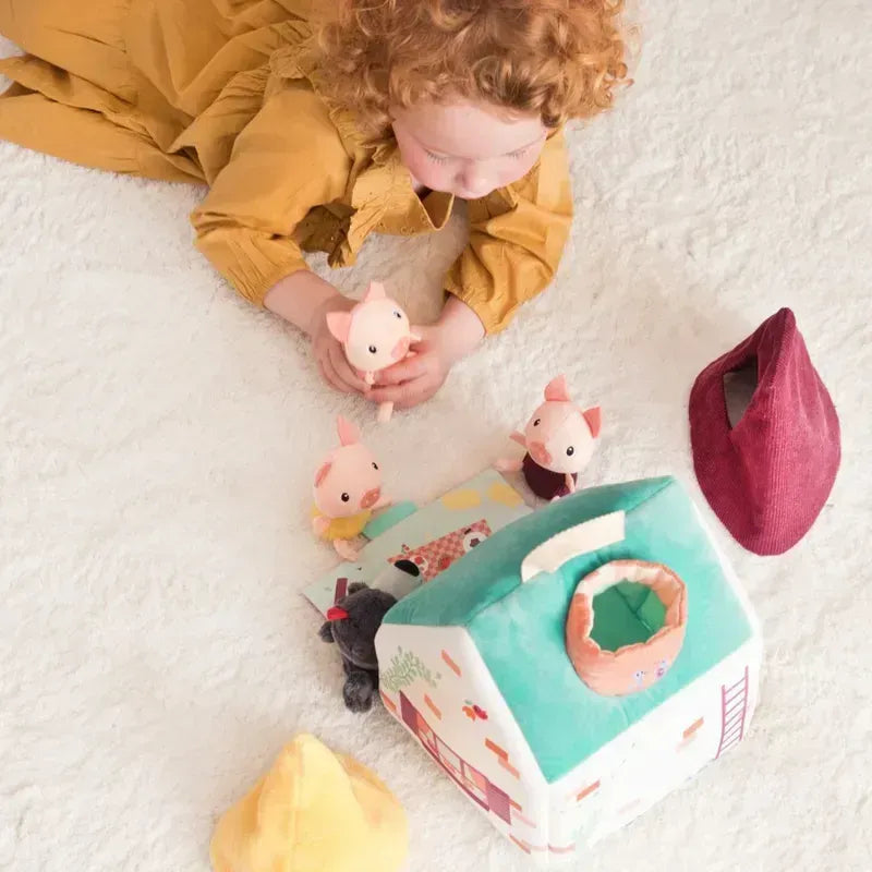 A young child with curly red hair wearing a mustard yellow dress is lying on a light-colored carpet. Engaged in creative play, the child is playing with the Lilliputiens Houses Of The Wolf And The 3 Little Pigs, enhancing fine motor skills. Other soft toys and accessories are scattered around.