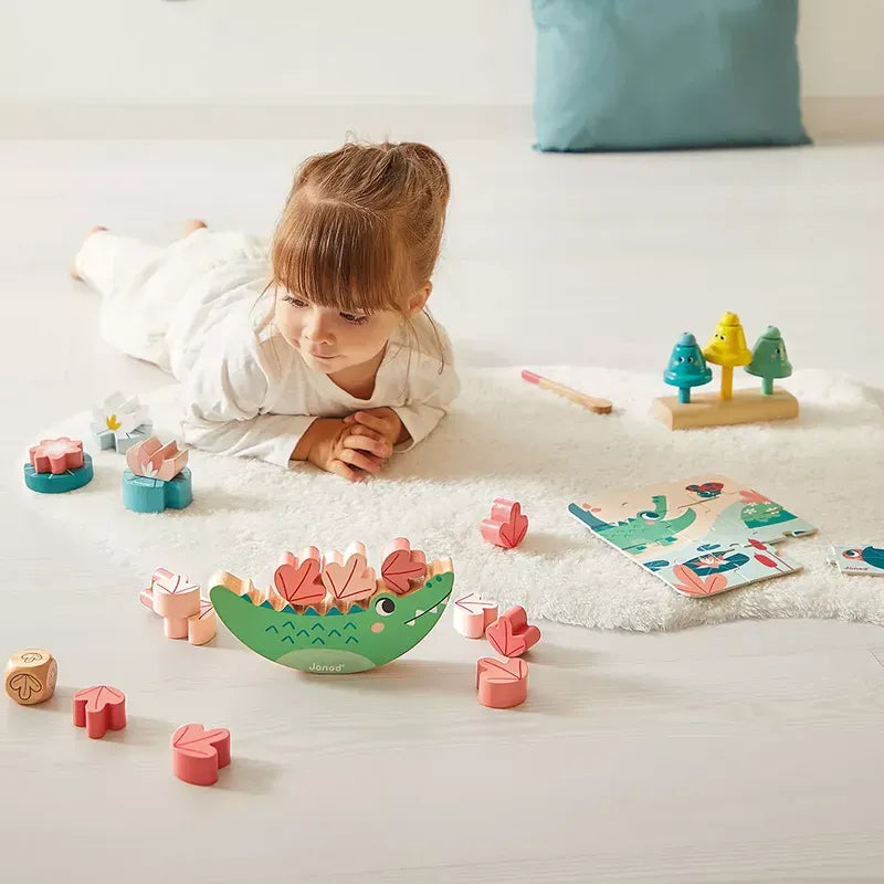 A child lays on the floor on their stomach, playing with colorful wooden blocks shaped like flowers and animals from the Janod Box of Toys 24 Months. These educational toys include a green piece with pink shapes stacked on top. Nearby, there are toy trees on a stand and an open book with illustrations, perfect for early-years development.