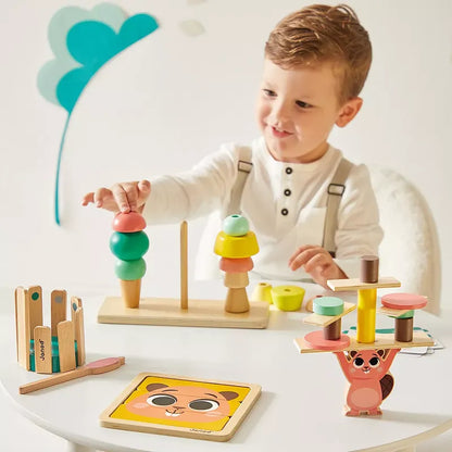 A young child with light brown hair and wearing an off-white shirt with suspenders sits at a table, engrossed in the Janod Box of Toys 36 Months. With sensory educational toys scattered around, the background is white adorned with playful wall decorations. The child looks focused and happy.