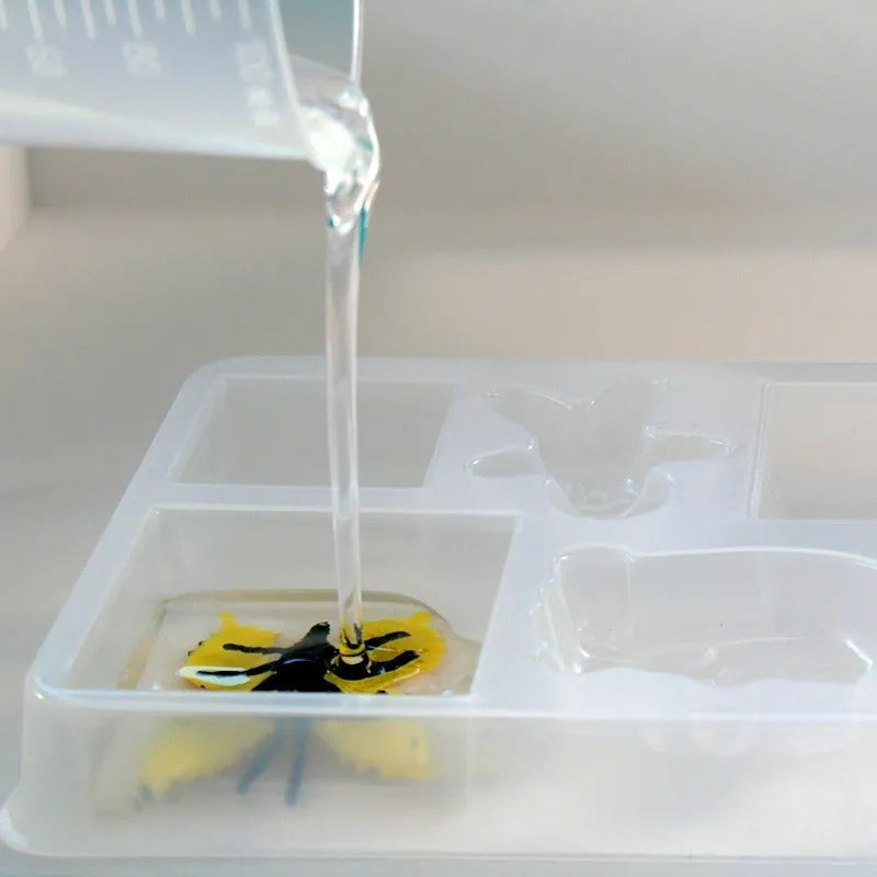 Clear liquid from a measuring cup is poured into a mold, capturing the butterfly's yellow and black details as part of the Sentosphere Resin Workshop DIY activity. The mold sits on a flat surface while the liquid fills the compartment, ready to hold decorative inclusions.