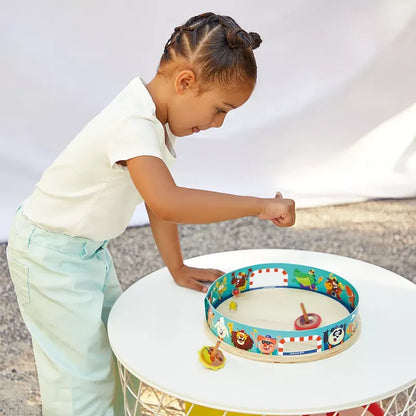 A child with braided hair, wearing a white shirt and light blue pants, stands next to a round white table. The child, aged 4 years and up, is playing with a Janod Applepop - Spinning Top Arena enclosed in a circular track featuring various cartoon animal characters.