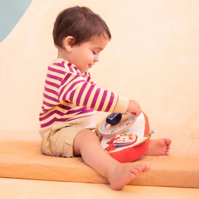 A toddler with short brown hair sits on a tan surface, wearing a striped long-sleeve shirt and beige shorts. The child is focused on a colorful Lilliputiens Farm Car Activity Panel featuring various shapes and textures, and the background is softly out of focus in warm tones.