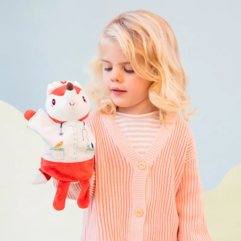 A young girl with blonde hair, wearing a pink cardigan and striped shirt, holds a Lilliputiens Alice Doctor Handpuppet. The puppet has white fur, red ears, and a red outfit with a stethoscope drawn on its coat. Perfect for imaginative play in a light blue and green background.