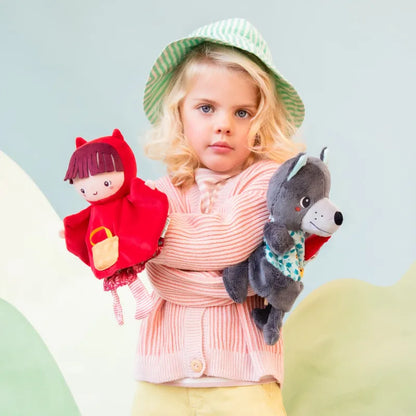 A young child with blonde hair, wearing a green and white striped hat and a peach-colored sweater, holds a Lilliputiens Red Riding Hood Handpuppet in one arm and a gray wolf plush toy in the other. The background is a soft, pastel-colored setting with abstract shapes, perfect for imaginative play.