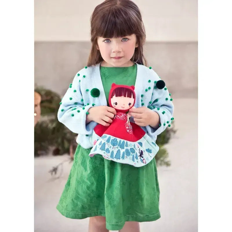 A young girl with long brown hair stands indoors holding the Lilliputiens Little Red Riding Hood Reversible Storydoll. The girl is wearing a green dress and a blue cardigan decorated with green pom-poms. The background is neutral with some greenery visible, lending an air of whimsical storytelling to the scene.