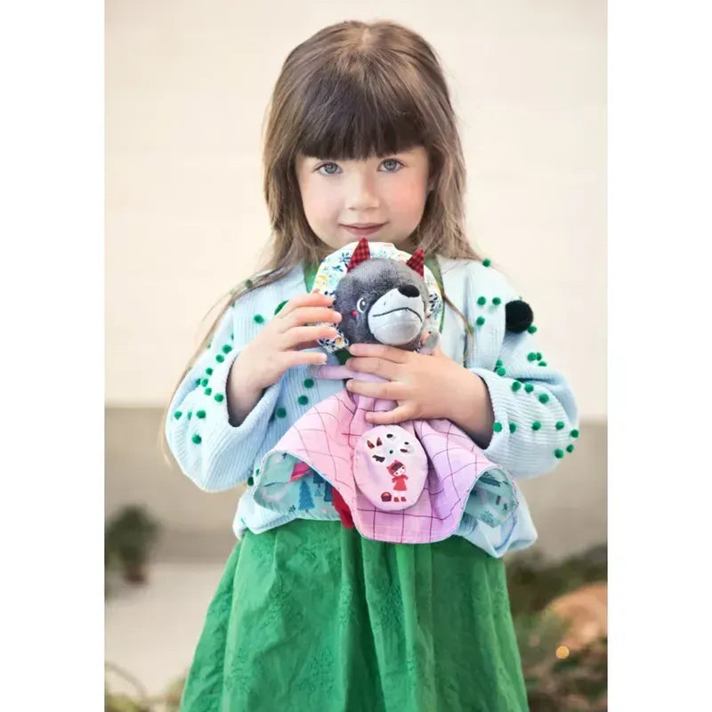 A young girl with long brown hair and bangs holds a Lilliputiens Little Red Riding Hood Reversible Storydoll. She is wearing a green dress and a white sweater with green dots. The background is blurred, with some green plants visible. She looks directly at the camera with a neutral expression.