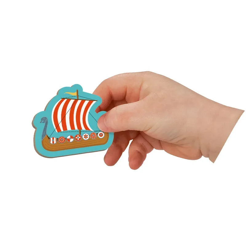 A hand holds the Janod History Magneti'book featuring a vibrant wooden Viking ship cutout, ideal for educational games. With its red and white striped sail, dragon-shaped prow, and ornate hull, it invites young adventurers to discover historical scenes against a plain white backdrop.