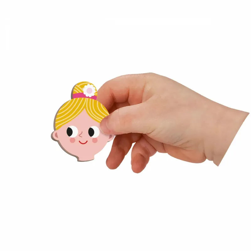 A hand holds a small, colorful cartoon cutout of a girl's head from the Janod Jobs Magneti'book, featuring a yellow hair bun with a pink flower. This educational toy showcases large eyes, a small smile, and simple facial design. The background is plain white.