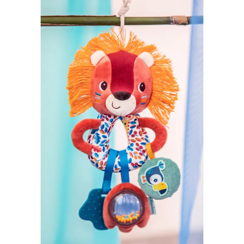 A hanging baby toy featuring a plush lion with a colorful mane and patterned body, ideal for enhancing motor and cognitive skills. It includes dangling blue and green shapes, a fish-themed baby's rattle, and a small plush bird. The Lilliputiens Jack Multi Activity Rings are suspended by a bamboo rod against a blurred blue and white background. Suitable for 3 months+.