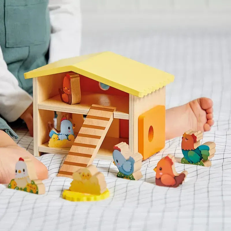 A child plays with the Janod Farm - My First Chicken Coop, a wooden early learning toy that includes a yellow-roofed house, a ramp, and several colorful wooden chicken figurines. The child's legs are visible on either side of this toddler educational toy, which is set on a white, textured surface.