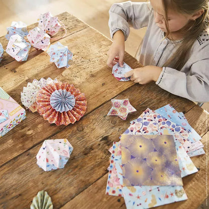 A young girl engages in the creative art of Janod Origami Delightful Decoration, carefully folding patterned paper from a creative kit to craft intricate designs, with a collection of completed colorful paper flowers scattered across a wooden table.
