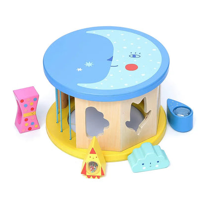 Introducing the Vilac Night and Day Shape Sorter, a colorful wooden baby toy featuring a moon and stars design on its blue lid. The sorter includes various shapes like a pink twisted block, a yellow and red rocket, a blue cloud, and a blue teardrop with a magnifying glass. This delightful toy is perfect for developing fine motor skills in young children.